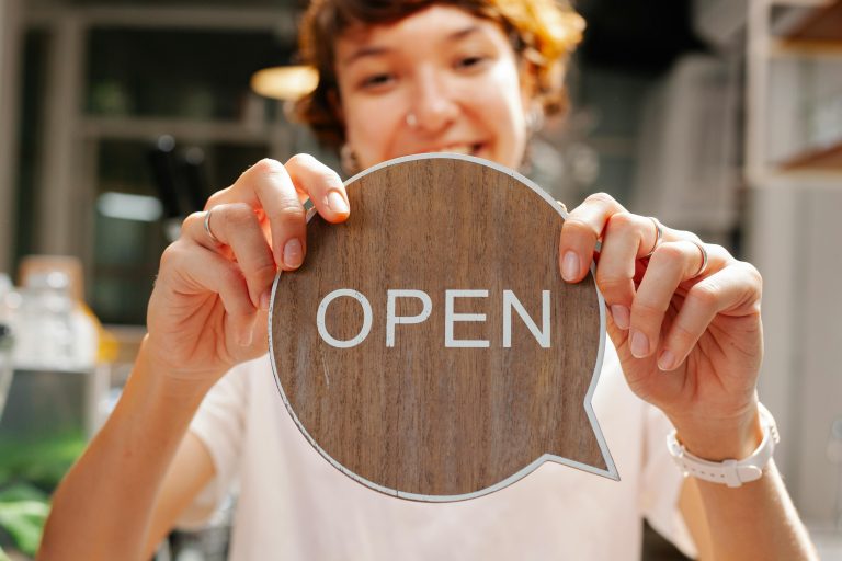 Happy young woman worker in casual clothes standing in light cafe and showing small sign with word open